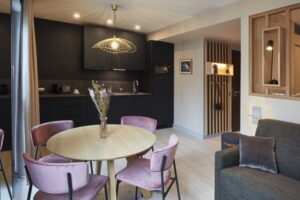 Wohnbereich eines Serviced Apartments mit schwarzer Küchenzeile, hellem Holztisch und rosa Polsterstühlen