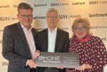 Zwei Männder und eine Frau präsentieren ein @home by Best Western-Schild