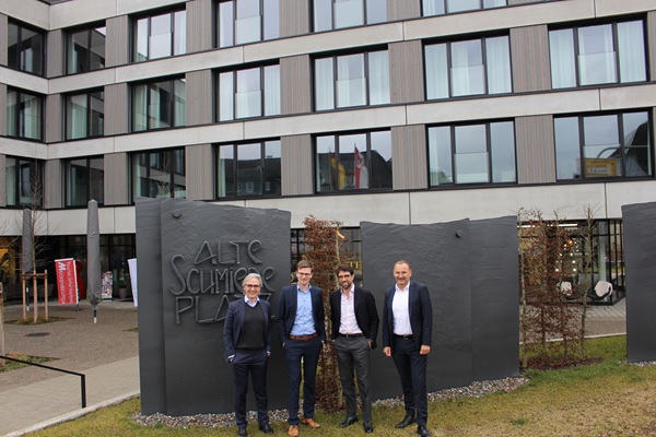 Vier Männer indunkler Kleidung vor einem modernen mehrgeschossigen Gebäude