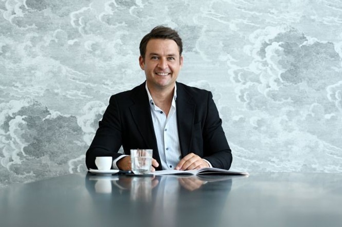 Smiling man in a dark blazer sits at a table with a notebook, glass of water, and coffee cup in front of him.