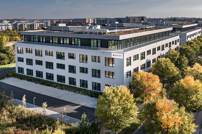 Modern white office building with many windows and a rooftop garden, surrounded by autumn trees.