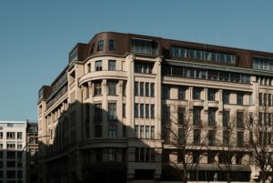 Large beige multi-story urban building with a curved corner, many windows, and leafless trees in the foreground.