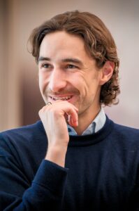Man with brown hair in a navy sweater smiles, resting his hand near his chin in a thoughtful pose.